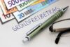 Handgeschriebener Hinweis „Grundfreibetrag“ neben Stift, Brille und Euro-Banknoten; Symbol für Einkommensteuer, Steuerfreibetrag, Steuern und private Finanzen.
