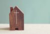 Kleines Holzhaus mit eingraviertem Kreuz vor pastellgrünem Hintergrund; Symbol für Kirche, Gemeinde, Glauben und Spenden.