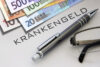 Krankengeldzuschuss bei stufenweiser Wiedereingliederung – Euro-Banknoten mit Stift und Brille auf Dokument