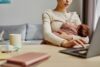 Frau hält ihr Baby im Arm und arbeitet dabei am Laptop an einem Tisch